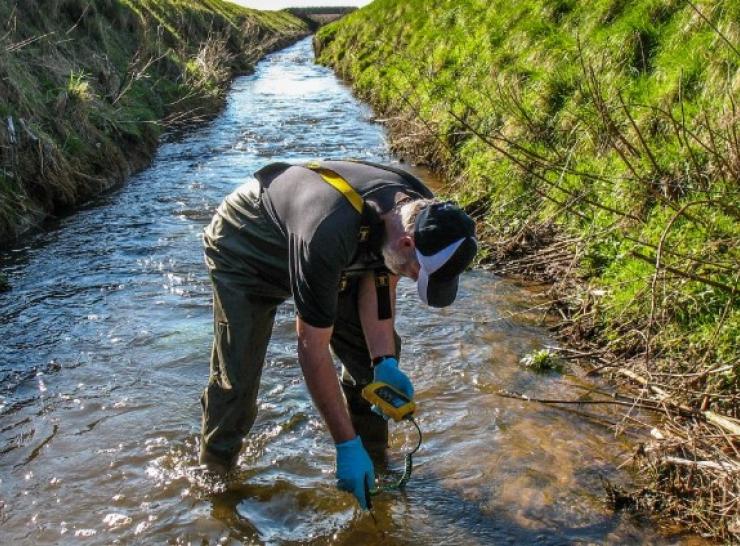 Person in waders and blue gloves bent in a shallow stream with grassy banks inserts a yellow monitoring device into the water